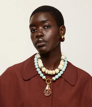 A woman with short hair wears a maroon coat and the Lizzie Fortunato Petra Collar—a large, layered necklace crafted from recycled white bone beads with gold-plated accents—along with a single oval earring, looking at the camera against a plain background.