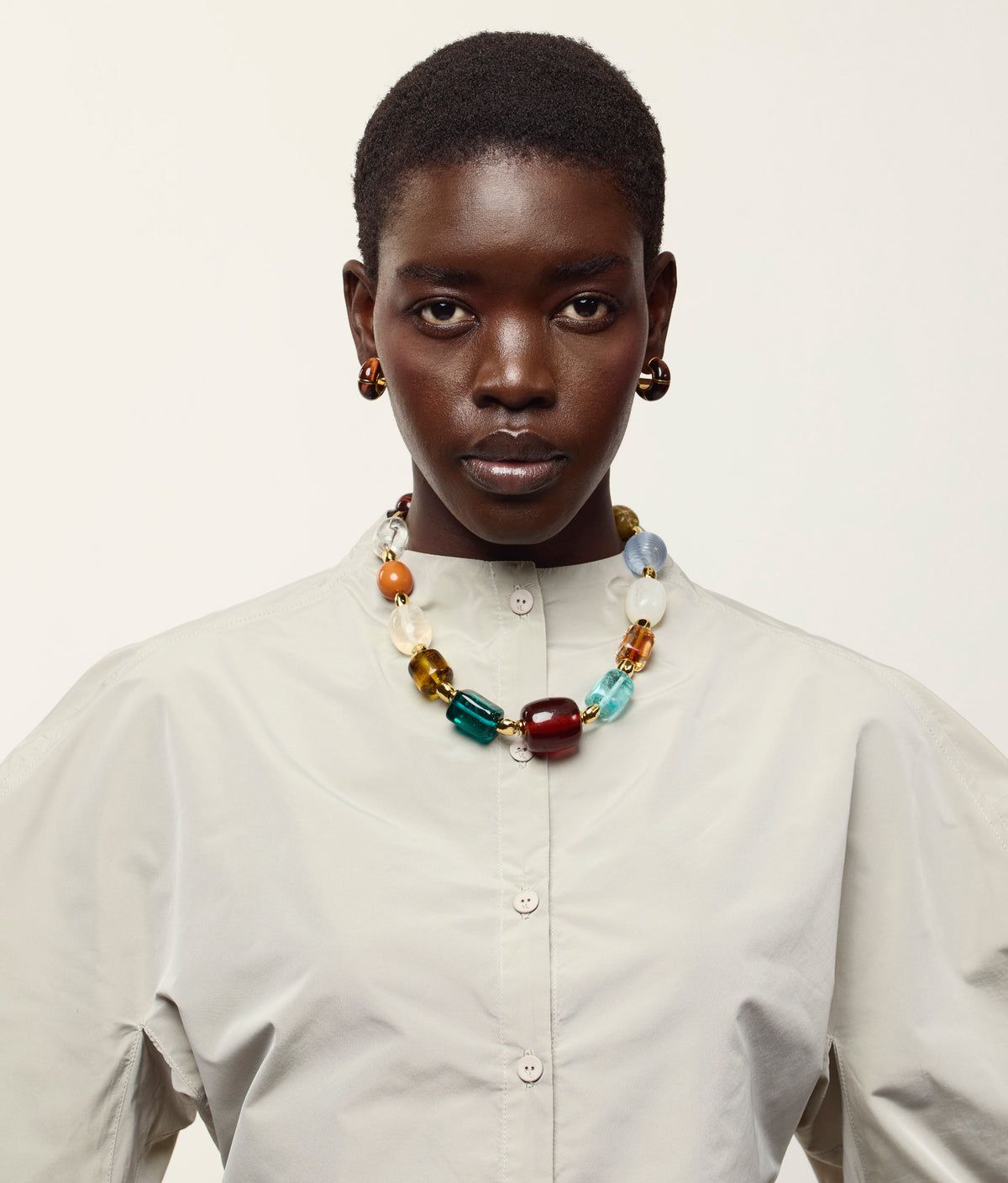 A person wearing a light-colored button-up shirt, the Lizzie Fortunato Acrobat Necklace, and hoop earrings, posing against a plain background.