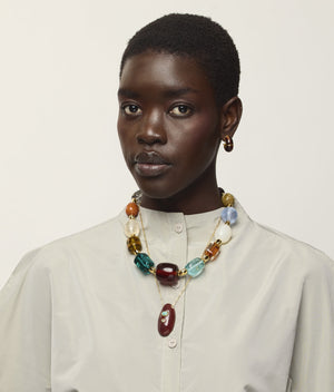 A woman with short hair wears a cream button-up shirt, gold hoop earrings, and the Lizzie Fortunato La Paz Pendant Necklace—a chunky, multicolored bead necklace featuring an amazonite ceramic pendant—set against a neutral background.