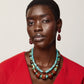 A person wearing a bright red top, the Lizzie Fortunato Granada Necklace featuring turquoise heishi beads and handmade porcelain accents, along with red drop earrings, stands against a plain white background.