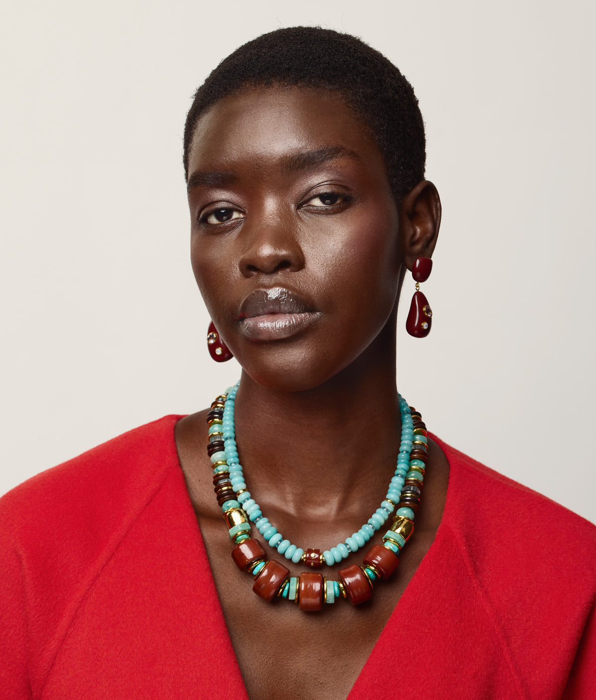 A person wearing a bright red top, the Lizzie Fortunato Granada Necklace featuring turquoise heishi beads and handmade porcelain accents, along with red drop earrings, stands against a plain white background.