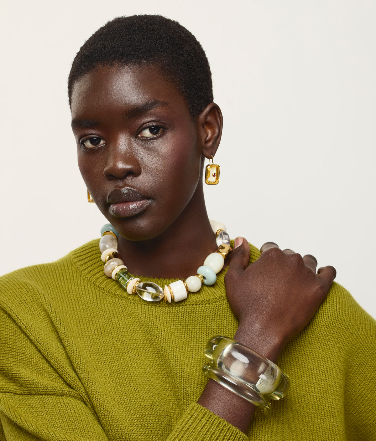 Woman wearing a green sweater, the Lizzie Fortunato Monument Collar necklace, a clear bracelet, and gold earrings poses against a plain white background.
