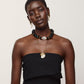 A woman with short hair wears a strapless black dress, gold earrings, and the Lizzie Fortunato Cirque Necklace—a chunky black and gold-plated beaded statement piece—layered with a delicate charm necklace, standing against a plain light background.