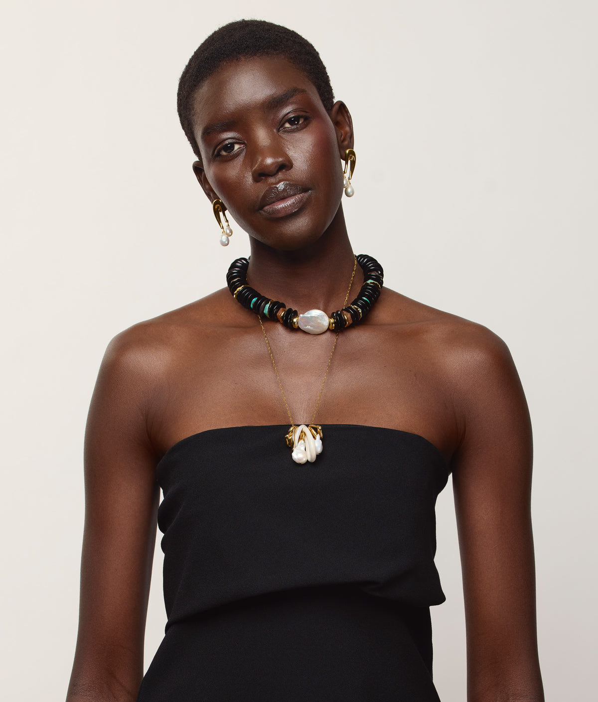 A woman with short hair wears a strapless black dress, gold earrings, and the Lizzie Fortunato Cirque Necklace—a chunky black and gold-plated beaded statement piece—layered with a delicate charm necklace, standing against a plain light background.