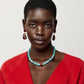 A woman with short hair wears a red top, the Lizzie Fortunato Hidden Stone Necklace featuring amazonite graduated beads and a brown accent bead, and red drop earrings, against a plain light background.