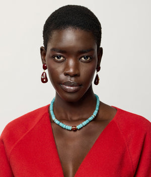 A woman with short hair wears a red top, the Lizzie Fortunato Hidden Stone Necklace featuring amazonite graduated beads and a brown accent bead, and red drop earrings, against a plain light background.
