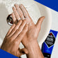 A person rubs a white exfoliating cream on their hands over a sink, with a blue bottle of Jack Black Face Buff Energizing Scrub placed nearby.