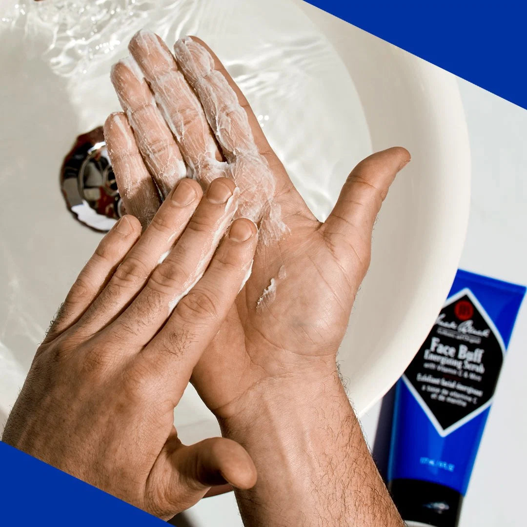 A person rubs a white exfoliating cream on their hands over a sink, with a blue bottle of Jack Black Face Buff Energizing Scrub placed nearby.