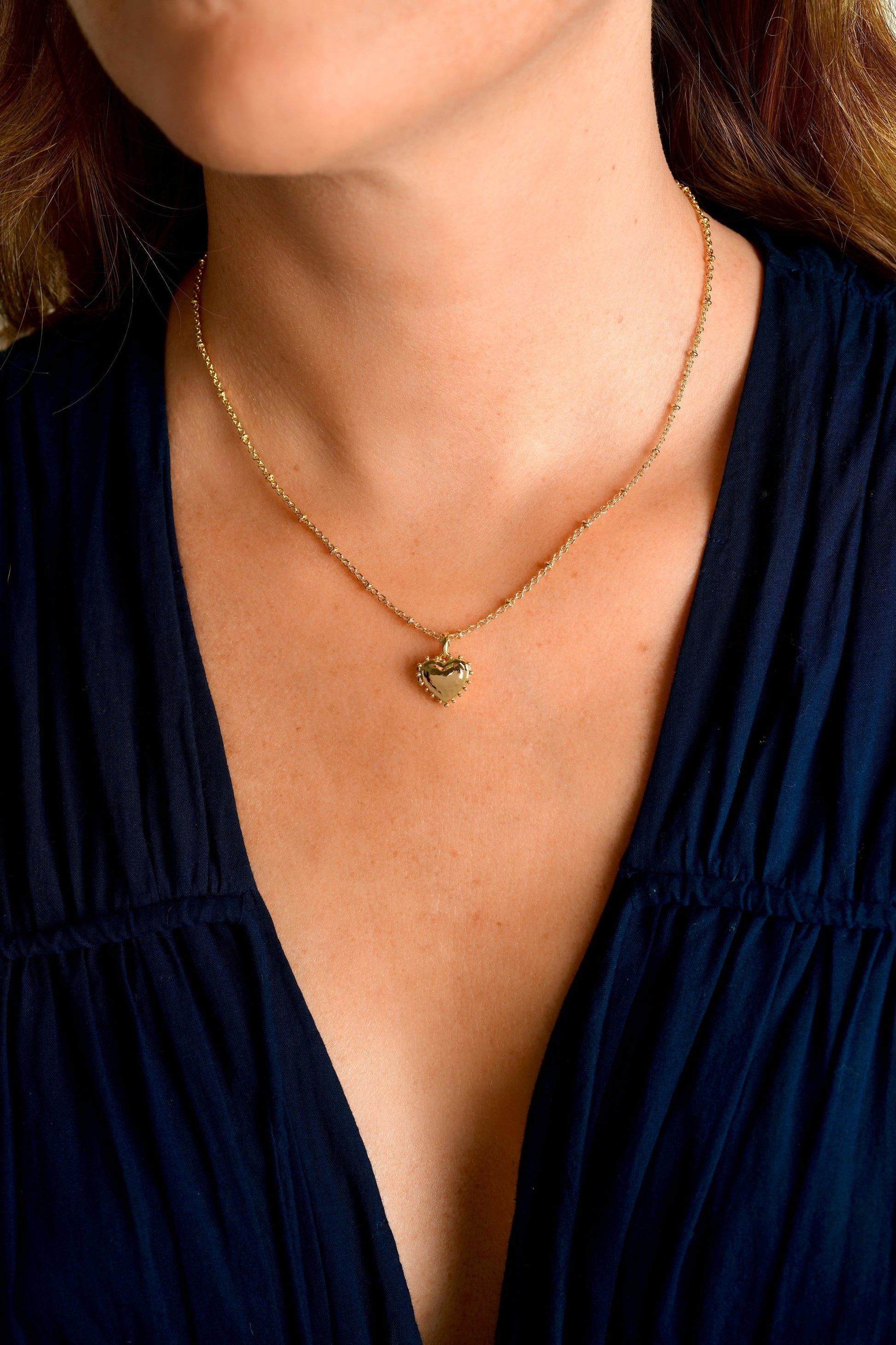 A person wearing the Anabel Aram Heart Locket Necklace, an 18k gold heart-shaped pendant with a scalloped edge, styled as keepsake jewelry and paired with a dark V-neck top.