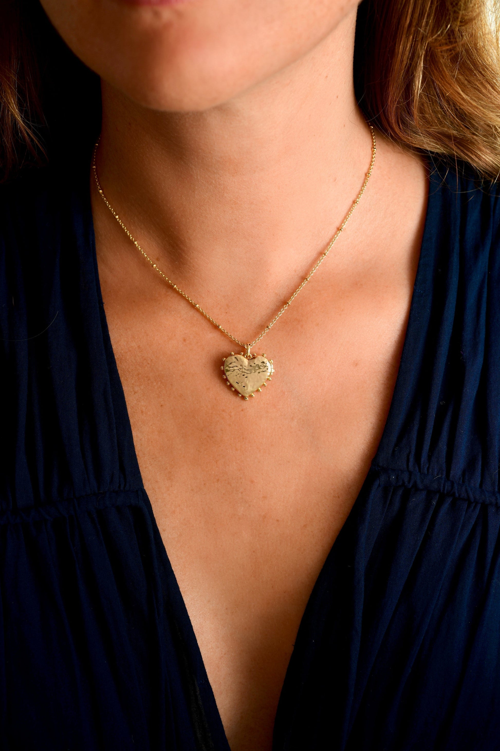 A person wears the Anabel Aram Heart Locket Necklace, a gold dipped brass piece, as a cherished keepsake with a dark V-neck top.