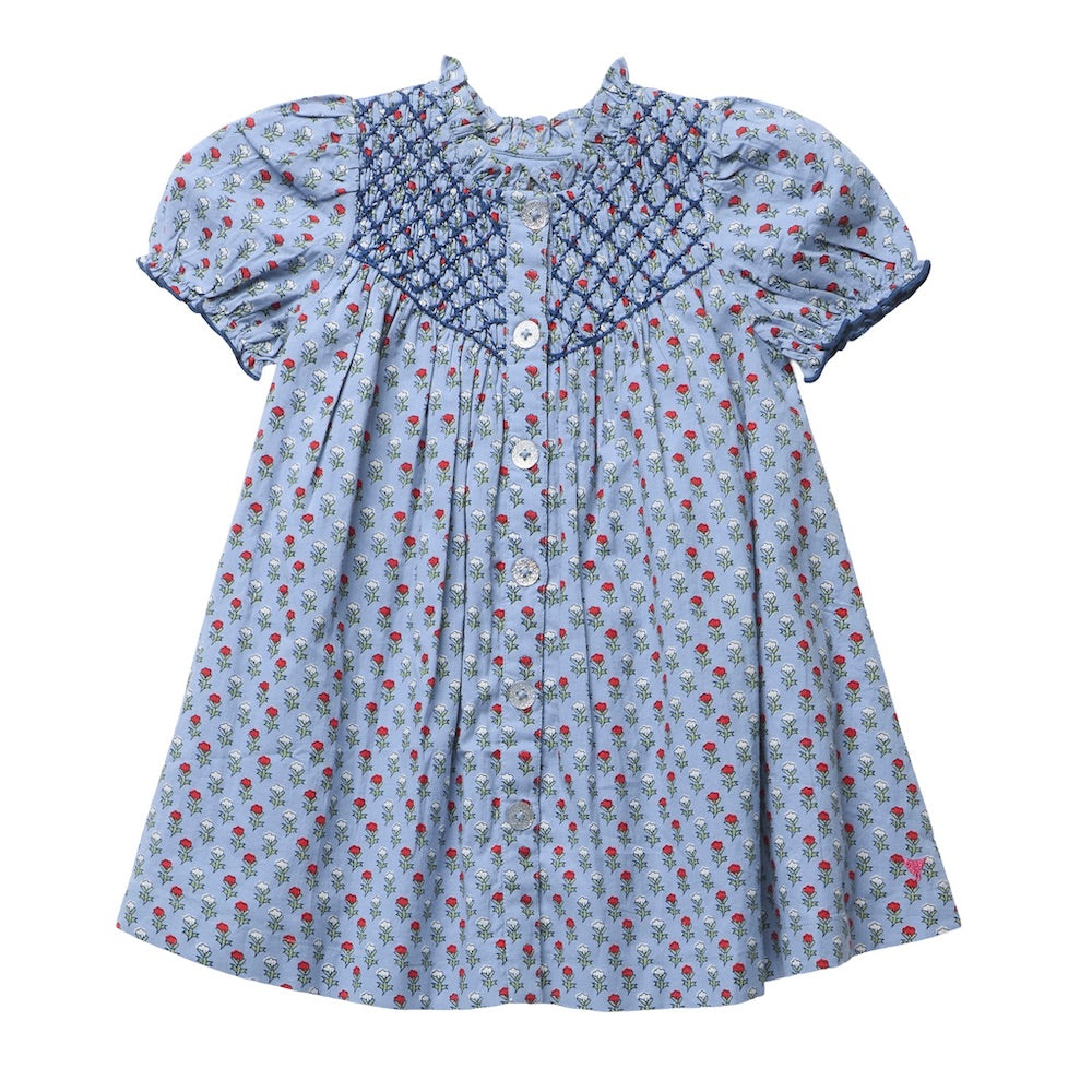 Girls’ Lucia Dress by Pink Chicken in light blue, featuring a red and green floral blockprint-inspired pattern, crisp white front buttons, and elegant navy smocking detail across the chest and sleeves.