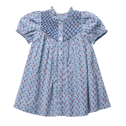 Girls’ Lucia Dress by Pink Chicken in light blue, featuring a red and green floral blockprint-inspired pattern, crisp white front buttons, and elegant navy smocking detail across the chest and sleeves.