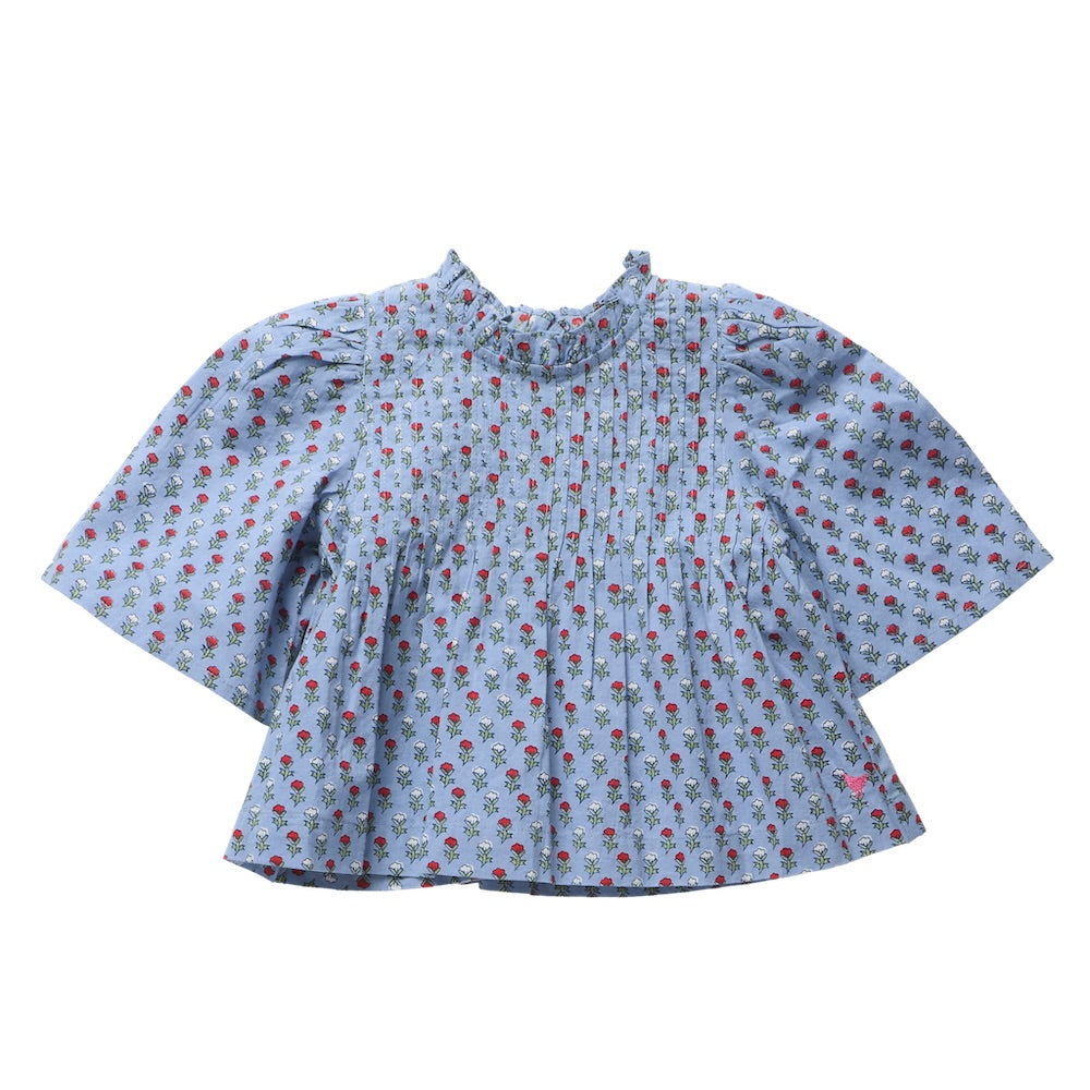 The Girls’ Jaipur Top by Pink Chicken features a light blue fabric adorned with a small red and green floral pattern, and is designed with a high collar, pleated front, wide bell sleeves, and finished with navy glitter buttons.