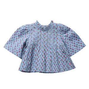The Girls’ Jaipur Top by Pink Chicken features a light blue fabric adorned with a small red and green floral pattern, and is designed with a high collar, pleated front, wide bell sleeves, and finished with navy glitter buttons.