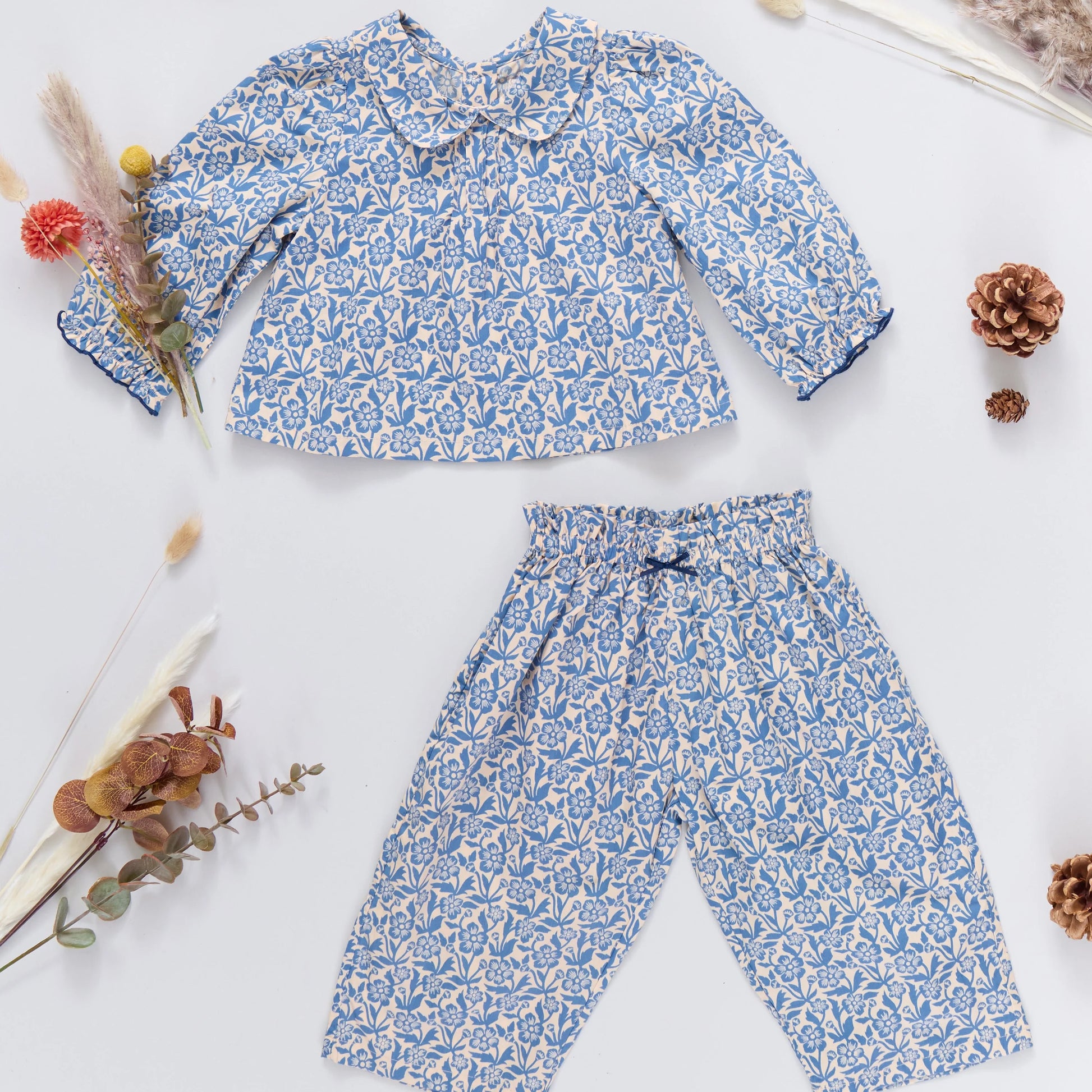 The Pink Chicken Baby Girls' Dawson 2-Piece Set includes a blue and white cotton poplin Climbing Floral Print long-sleeve shirt and matching pants, styled with dried flowers and pinecones on a white background.