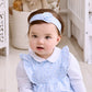 A baby wearing the Kissy Kissy Fall in Bloom Headband sits indoors on a light-colored floor, with baskets and stuffed animals in the background.