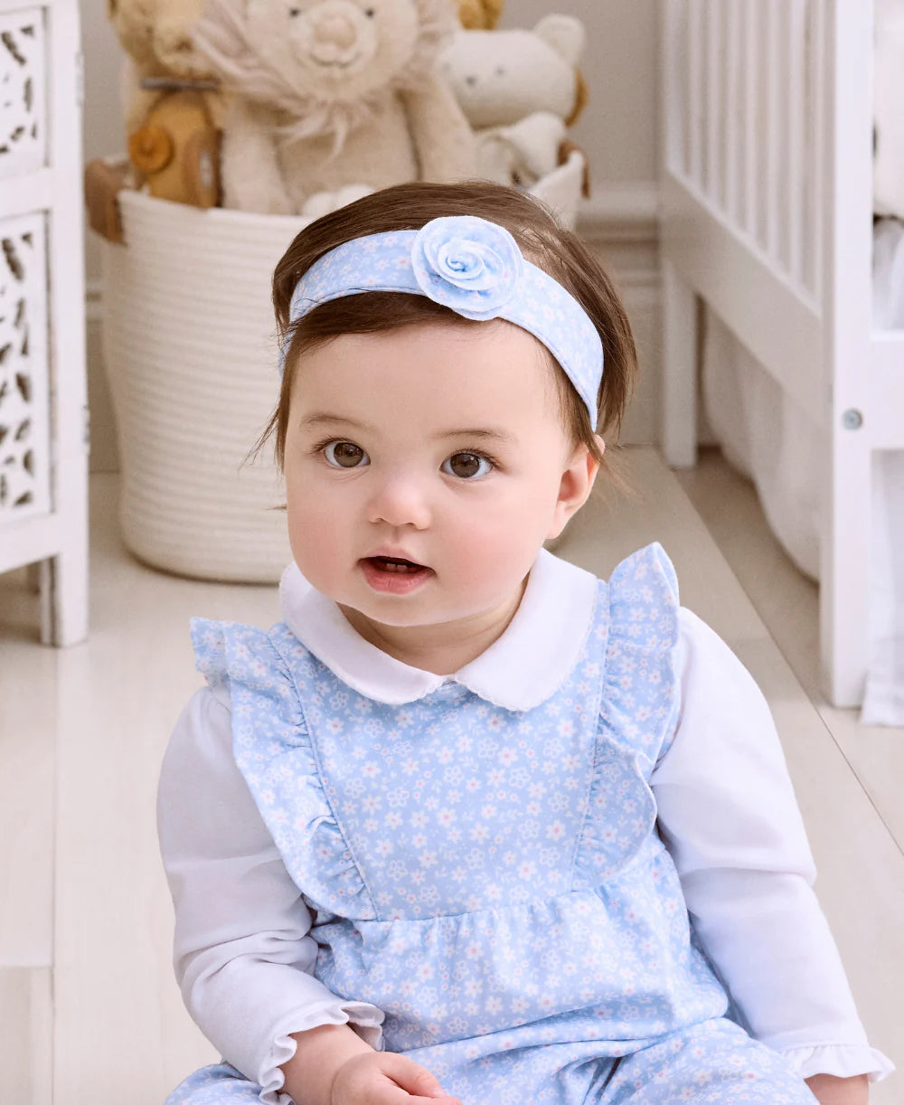 A baby wearing the Kissy Kissy Fall in Bloom Headband sits indoors on a light-colored floor, with baskets and stuffed animals in the background.