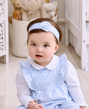 A baby wearing the Kissy Kissy Fall in Bloom Headband sits indoors on a light-colored floor, with baskets and stuffed animals in the background.
