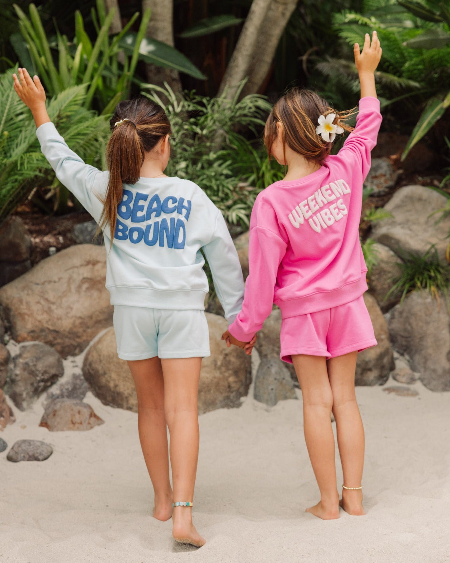Two girls stand on the sand holding hands, both wearing matching Feather 4 Arrow Baby Girls’ and Girls’ Weekend Sweatshirts, featuring trendy puff print graphics that read “Beach Bound” and “Weekend Vibes,” with their arms raised amidst rocks and tropical plants.