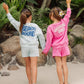 Two girls stand on sand, holding hands with their backs to the camera. One wears a blue Feather 4 Arrow “BEACH BOUND” Weekend Short set made of comfy French terry sweatshorts featuring an elastic waistband and drawcord, while the other sports a pink Feather 4 Arrow “WEEKEND VIBES” Weekend Short set. Tropical plants can be seen in the background.