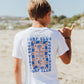 A boy stands on a sandy beach, facing away, wearing the Feather 4 Arrow Boys’ Surf Club Good Vibes Vintage Tee crafted from premium cotton and flashing a shaka hand gesture. The blurred shoreline and a blue sky fill the background.