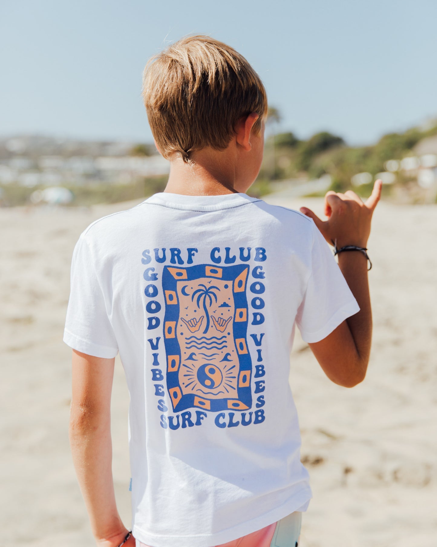A boy stands on a sandy beach, facing away, wearing the Feather 4 Arrow Boys’ Surf Club Good Vibes Vintage Tee crafted from premium cotton and flashing a shaka hand gesture. The blurred shoreline and a blue sky fill the background.