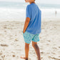 Wearing the Feather 4 Arrow Baby Boys’ and Boys’ High Tide Pocket Tee, a boy walks barefoot on a sandy beach in patterned shorts, with the ocean and another person visible in the background.