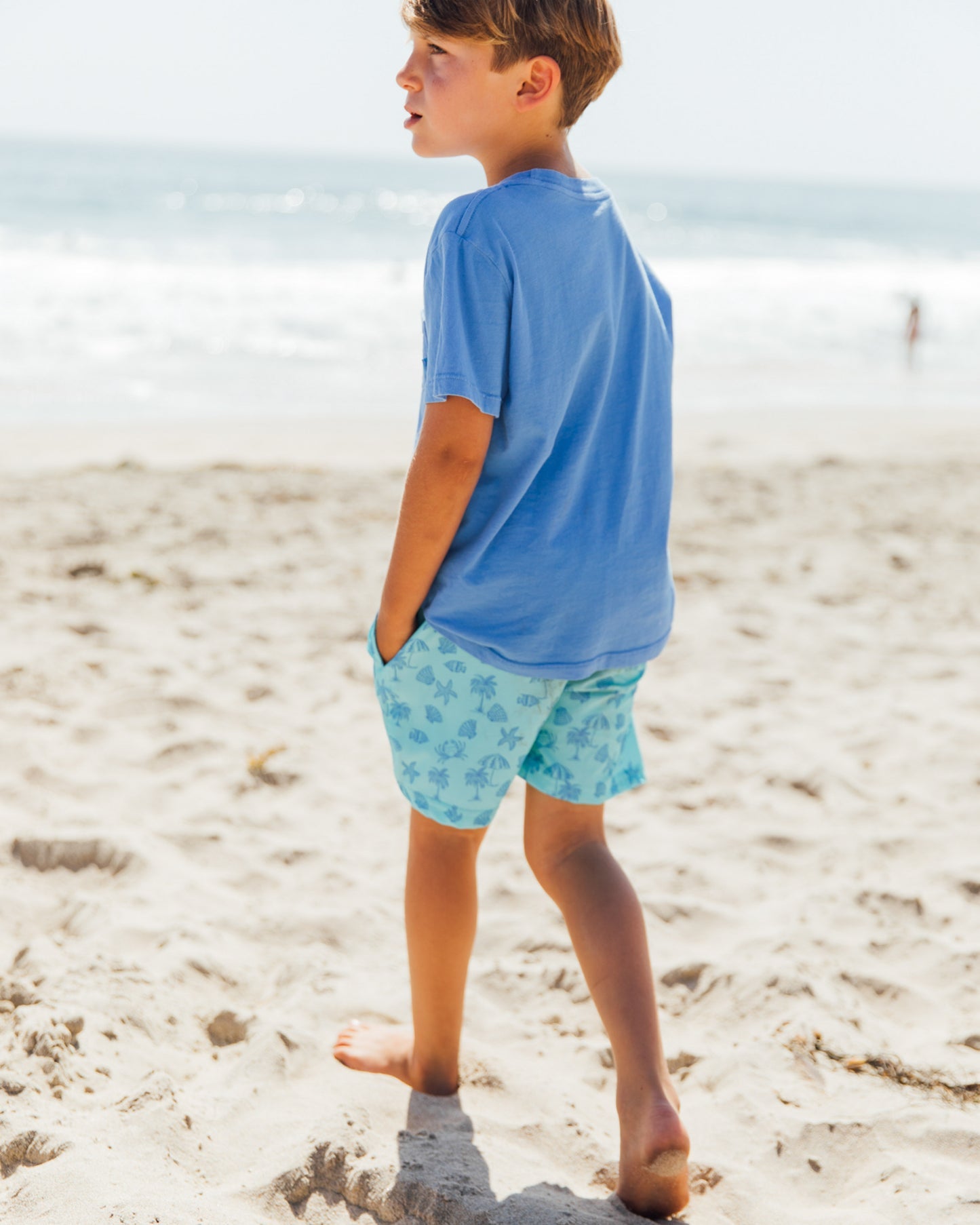 Wearing the Feather 4 Arrow Baby Boys’ and Boys’ High Tide Pocket Tee, a boy walks barefoot on a sandy beach in patterned shorts, with the ocean and another person visible in the background.