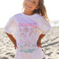 A smiling child stands on a sandy beach wearing the Feather 4 Arrow Girls’ Oversized Tee, crafted from premium cotton and adorned with pastel seashell designs alongside the words “Seashells by the seashore.”.
