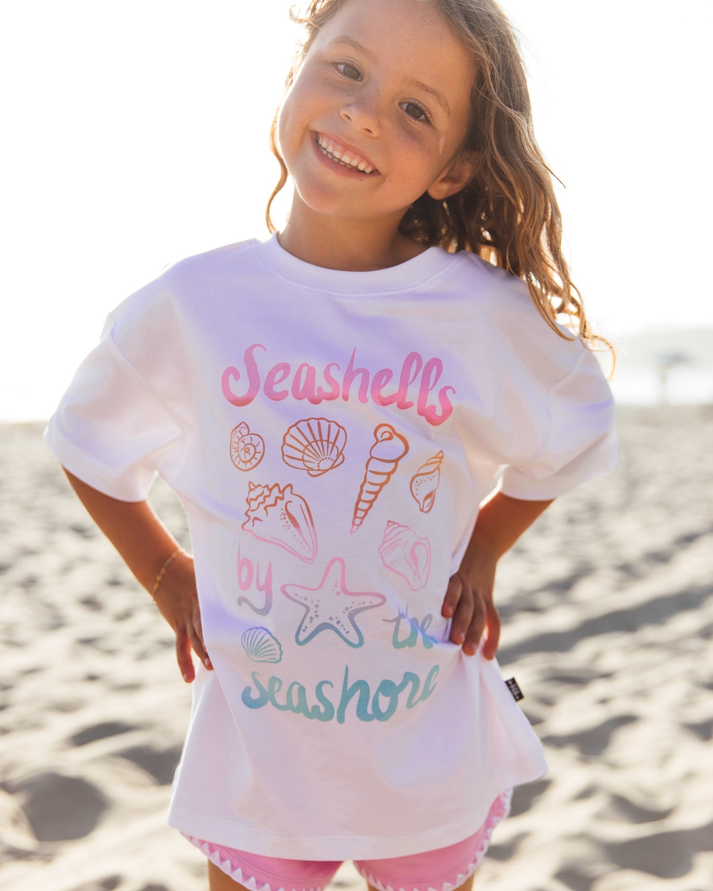 A smiling child stands on a sandy beach wearing the Feather 4 Arrow Girls’ Oversized Tee, crafted from premium cotton and adorned with pastel seashell designs alongside the words “Seashells by the seashore.”.