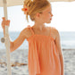 A girl in the Feather 4 Arrow Girls’ Swing Tank with a peach striped, flowy fit stands under a beach umbrella, holding the pole, with the ocean and sand in the background.
