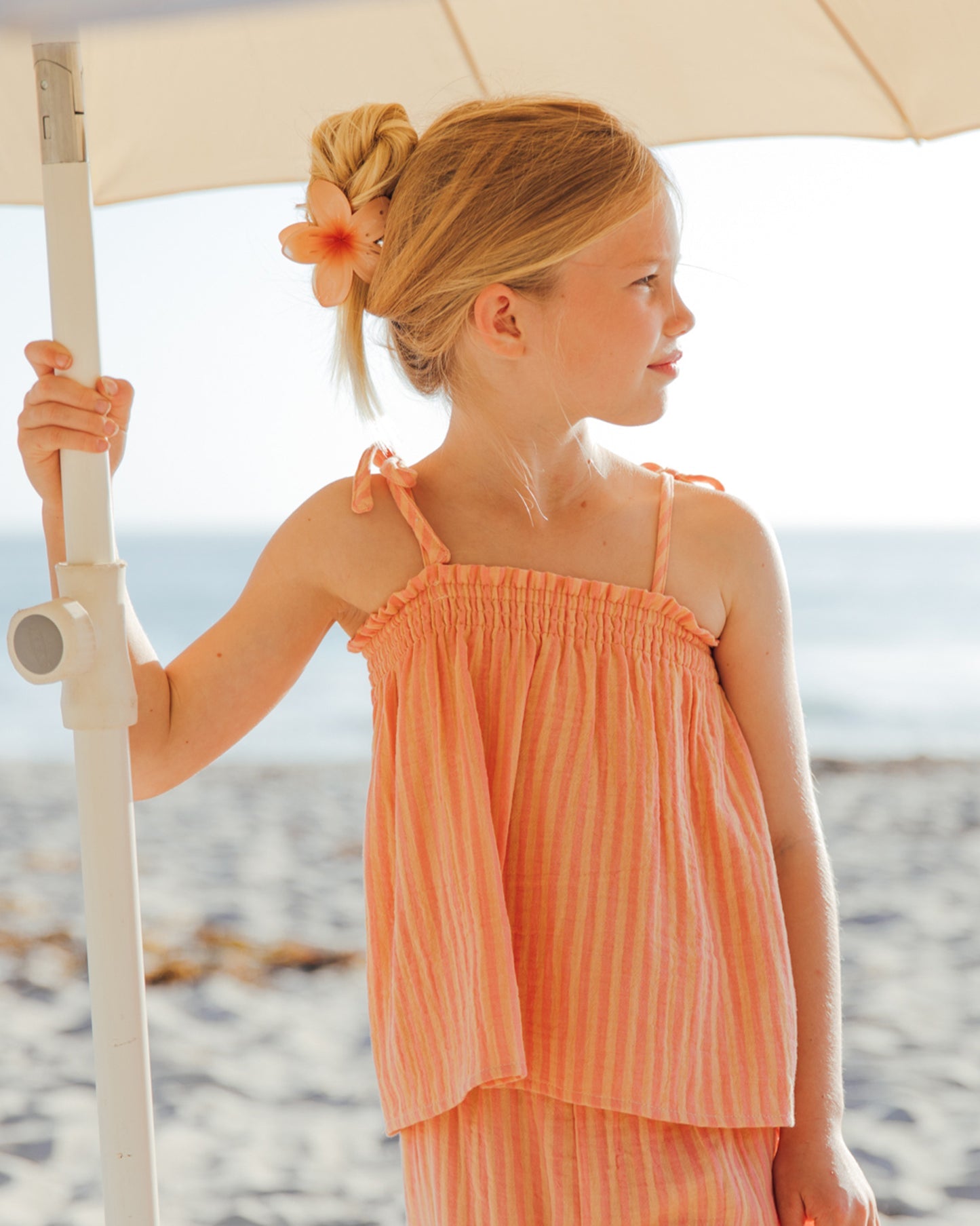 A girl in the Feather 4 Arrow Girls’ Swing Tank with a peach striped, flowy fit stands under a beach umbrella, holding the pole, with the ocean and sand in the background.