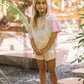 A young girl stands outdoors on stepping stones, dressed in the Feather 4 Arrow Girls’ Terry Hoodie and matching shorts in light pink, surrounded by greenery and wooden structures.