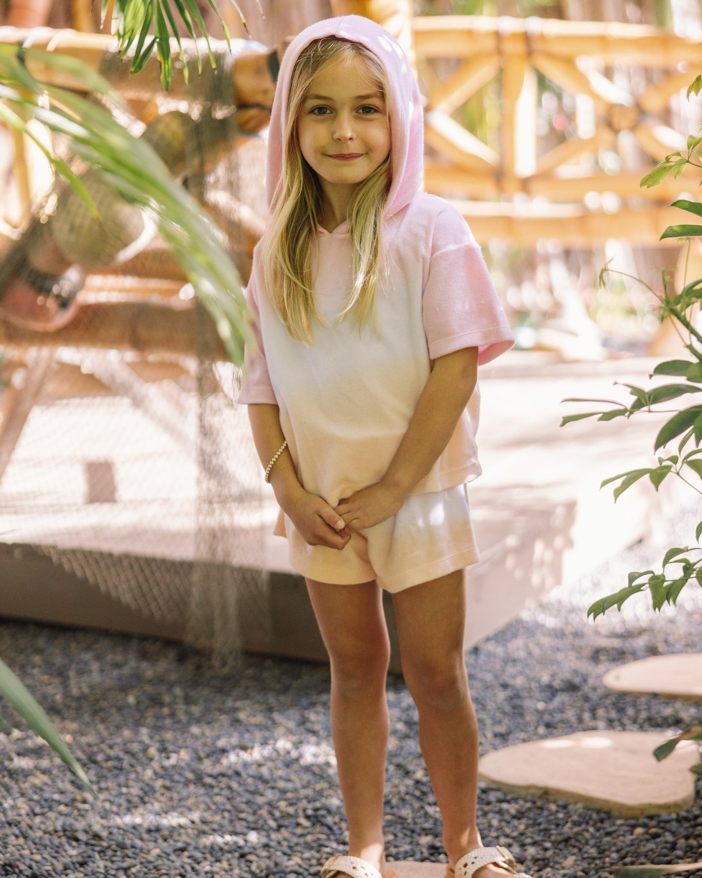 A young child stands outdoors on a stone path surrounded by greenery, looking at the camera while wearing Feather 4 Arrow’s Girls’ Weekend Short paired with a pink hooded shirt.