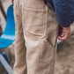 A close-up of a person wearing Feather 4 Arrow’s Boys’ Carpenter Twill Pant in tan, featuring durable cotton twill fabric and practical carpenter pockets, paired with a blue textured sweater while standing next to a blue playground seat outdoors.