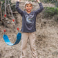 A young boy stands on a slackline while holding a metal bar above, next to a blue swing, wearing Feather 4 Arrow Boys’ Carpenter Twill Pants, with carpenter pockets for durability, with a dirt hillside and greenery in the background.