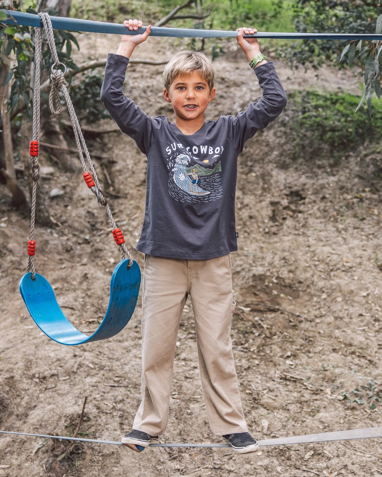 A young boy stands on a slackline while holding a metal bar above, next to a blue swing, wearing Feather 4 Arrow Boys’ Carpenter Twill Pants, with carpenter pockets for durability, with a dirt hillside and greenery in the background.