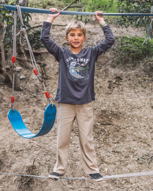 A young boy stands on a slackline while holding a metal bar above, next to a blue swing, wearing Feather 4 Arrow Boys’ Carpenter Twill Pants, with carpenter pockets for durability, with a dirt hillside and greenery in the background.