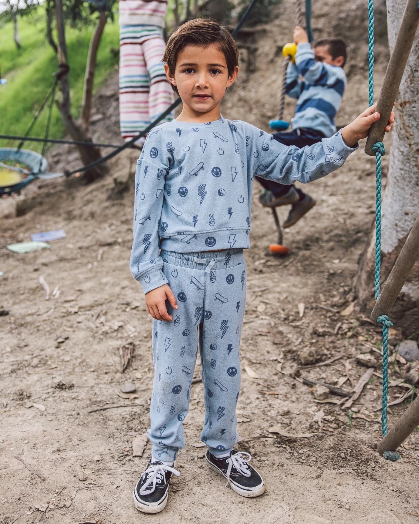 A young boy wears the Feather 4 Arrow Baby Boys’ and Boys’ Icon Hacci Set—crafted from ultra-soft hacci fabric—as he stands outdoors on the dirt, while another child enjoys climbing ropes in the background. This set is perfect kids loungewear for playtime adventures.