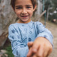 A smiling young boy in the Feather 4 Arrow Baby Boys’ and Boys’ Icon Hacci Set stands outdoors, reaching out toward the camera with both hands, showing off his comfy loungewear top and bottom made from soft hacci fabric.