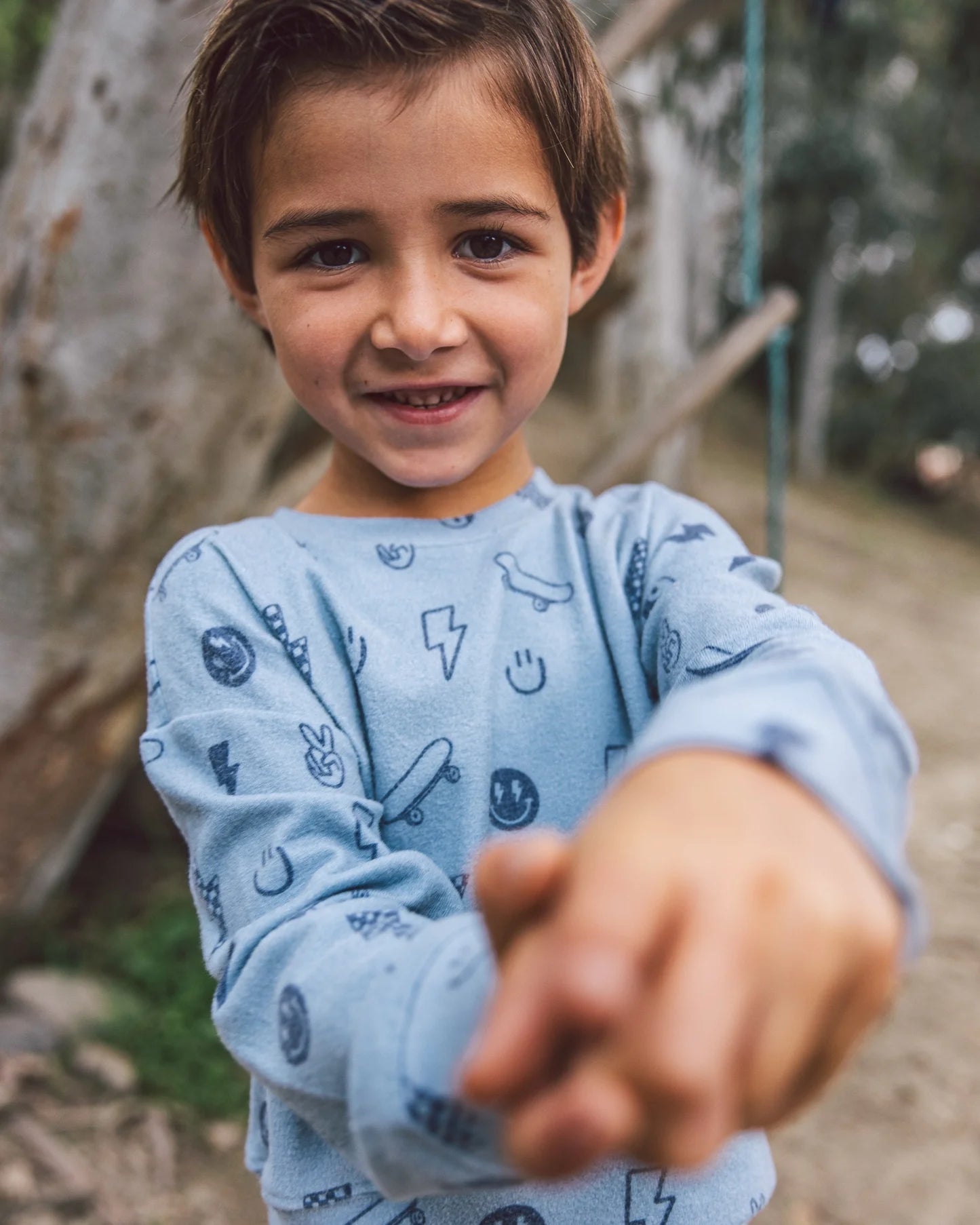 A smiling young boy in the Feather 4 Arrow Baby Boys’ and Boys’ Icon Hacci Set stands outdoors, reaching out toward the camera with both hands, showing off his comfy loungewear top and bottom made from soft hacci fabric.