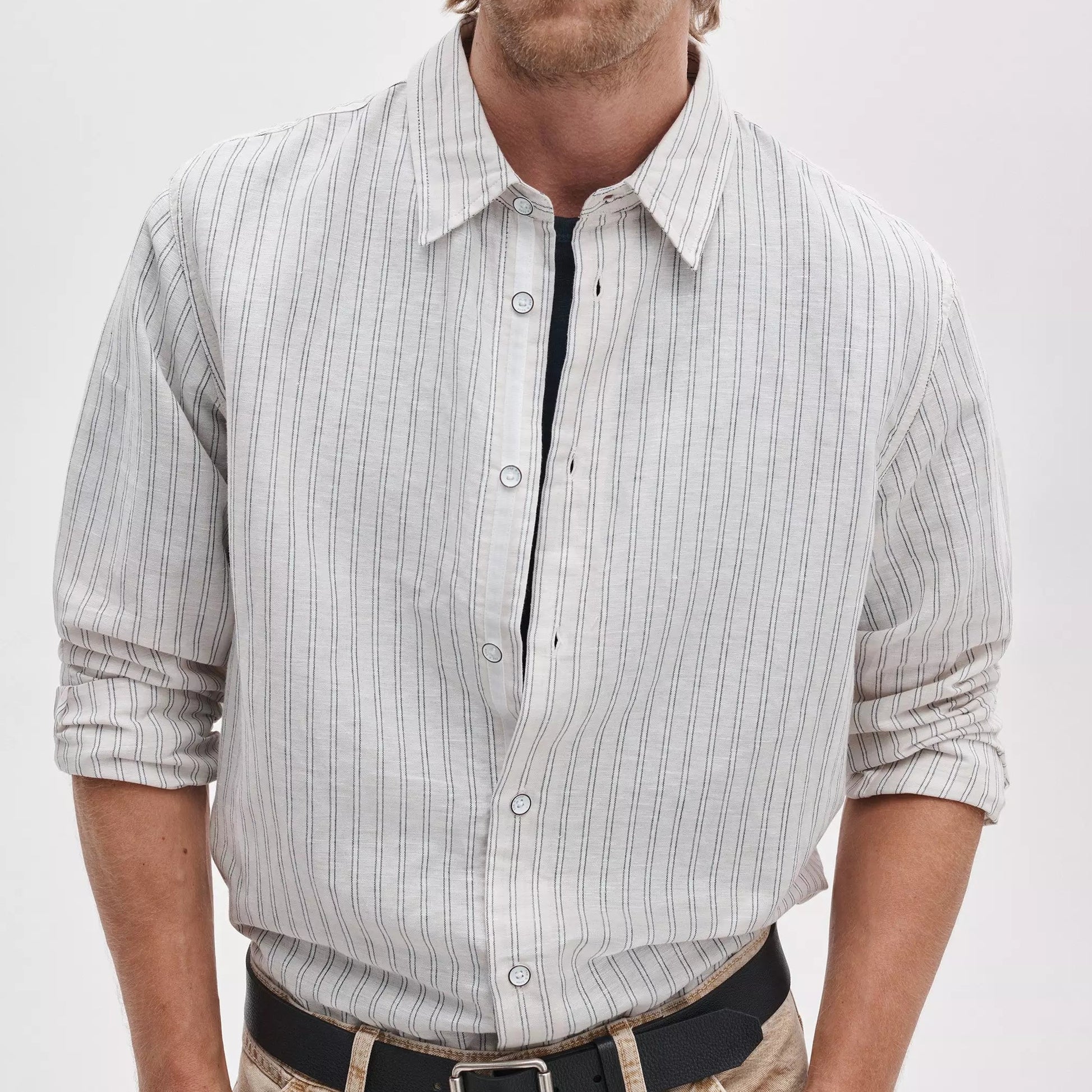 A man facing forward wears the Rag & Bone Finch Linen Stripe Shirt—a light-colored, relaxed-fit button-up featuring thin vertical stripes—paired with tan pants and rolled-up sleeves.