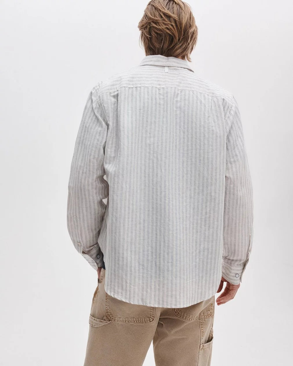 A person with light brown hair is wearing the Rag & Bone Finch Linen Stripe Shirt—a relaxed fit, button-up striped shirt—paired with beige pants, standing facing away from the camera against a plain white background.