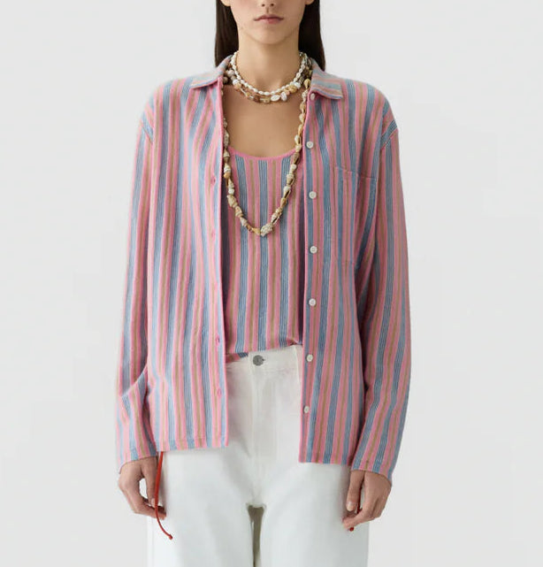A woman wears the Fine Stripe Showtime Shirt by Guest in Residence—a relaxed fit pink, blue, and white stripe button-down—over a matching top with white pants and layered shell necklaces, posed against a plain background.