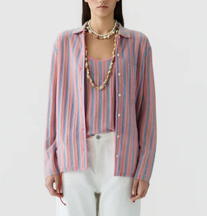 A woman wears the Fine Stripe Showtime Shirt by Guest in Residence—a relaxed fit pink, blue, and white stripe button-down—over a matching top with white pants and layered shell necklaces, posed against a plain background.