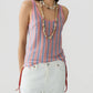 A person in the Guest in Residence Fine Stripe Tank, featuring pink, blue, and white stripes, styled with white pants with red side ties and layered shell necklaces, stands against a plain light background.