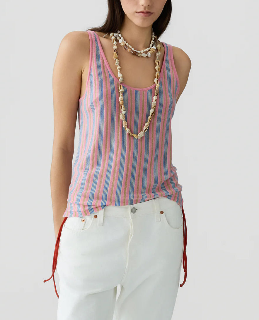 A person in the Guest in Residence Fine Stripe Tank, featuring pink, blue, and white stripes, styled with white pants with red side ties and layered shell necklaces, stands against a plain light background.