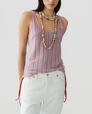 A person in the Guest in Residence Fine Stripe Tank, featuring pink, blue, and white stripes, styled with white pants with red side ties and layered shell necklaces, stands against a plain light background.