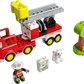 The LEGO® Duplo Fire Truck set by LEGO® features a fire truck with a ladder, firefighter and white cat figures, a drawing tile, and a green tree, all displayed on a white background.