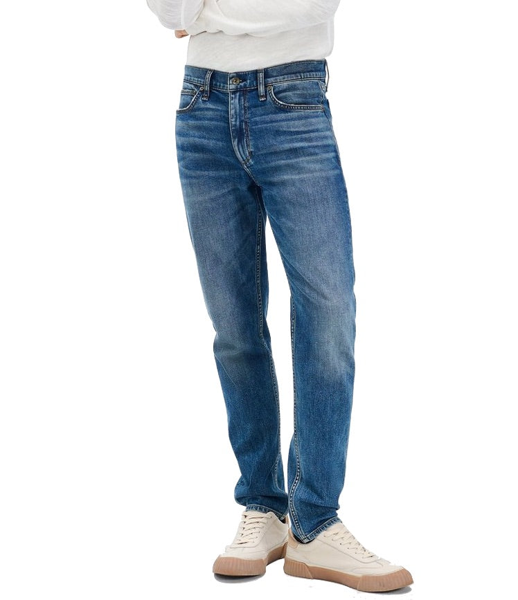 A person wearing Rag & Bone Fit 2 Authentic Stretch Denim jeans in medium indigo, paired with a white shirt and off-white sneakers, stands facing forward with arms folded.